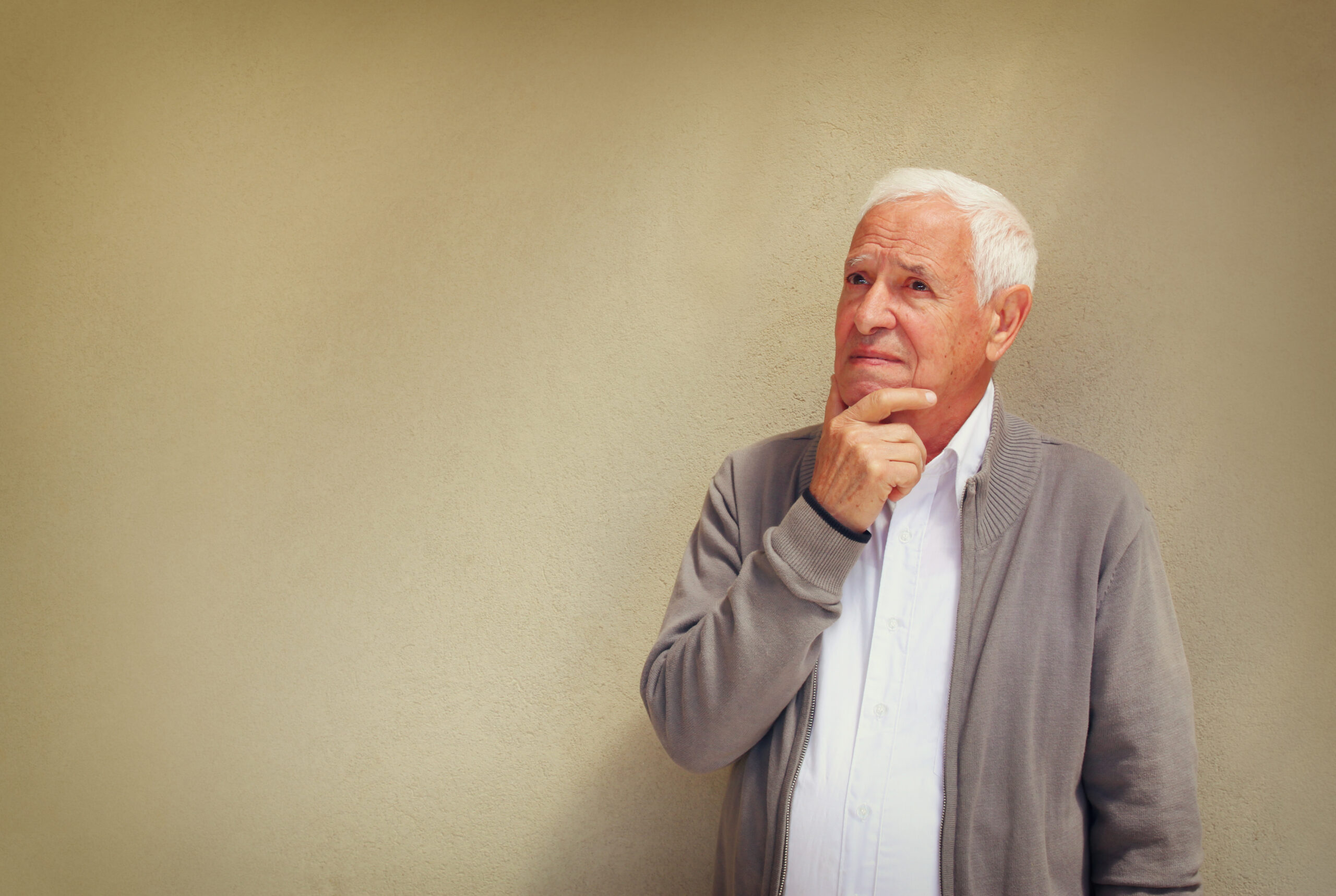 older man standing in front of tan wall while holding his chin